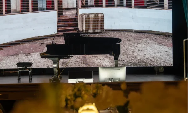 Pianoforte a coda nero sul palcoscenico davanti a maxischermo con allestimento floreale sfocato in primo piano